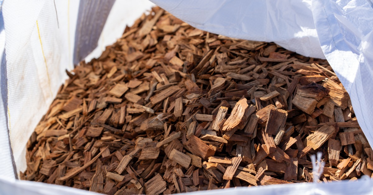 A close-up view of brown wood chips and mulch filling a large white bulk bag outdoors in bright sunlight.