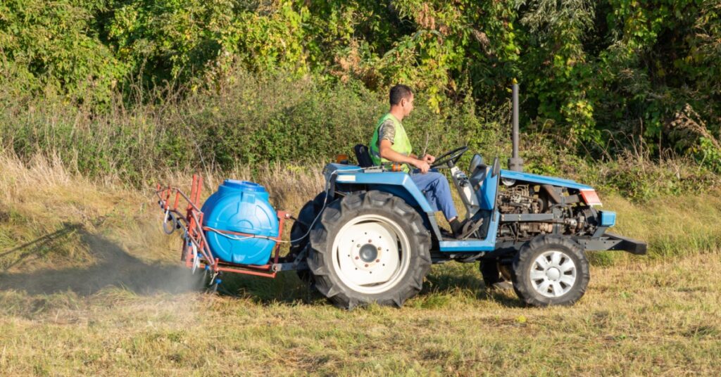 A man driving a blue tractor with a spray tank through dry grass beside dense green trees on a sunny day.